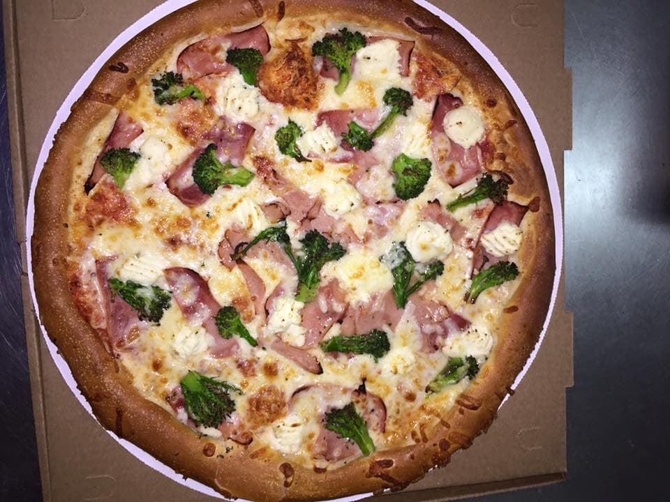 Freshly baked pizza with ham, broccoli, and white cheese dollops in a cardboard box.